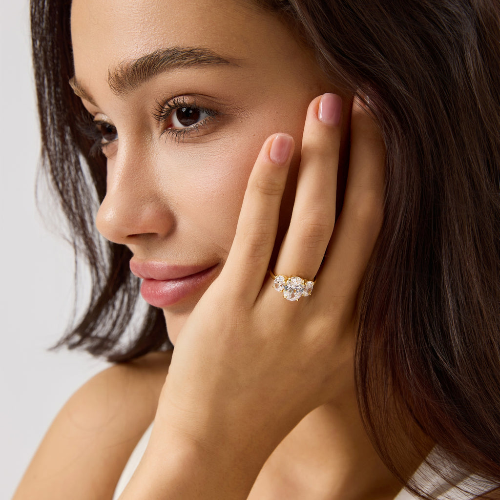 Woman wearing gold oval three stone engagement ring, touching her face.